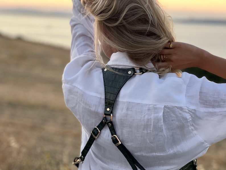 May include: A green leather camera strap with gold hardware. The strap is worn over the shoulders and has a criss-cross design in the back.