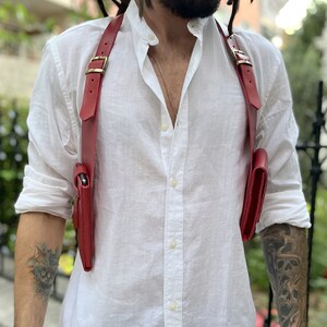 May include: A red leather crossbody bag with two separate compartments. The bag is worn over the shoulder and has a strap that goes across the chest.