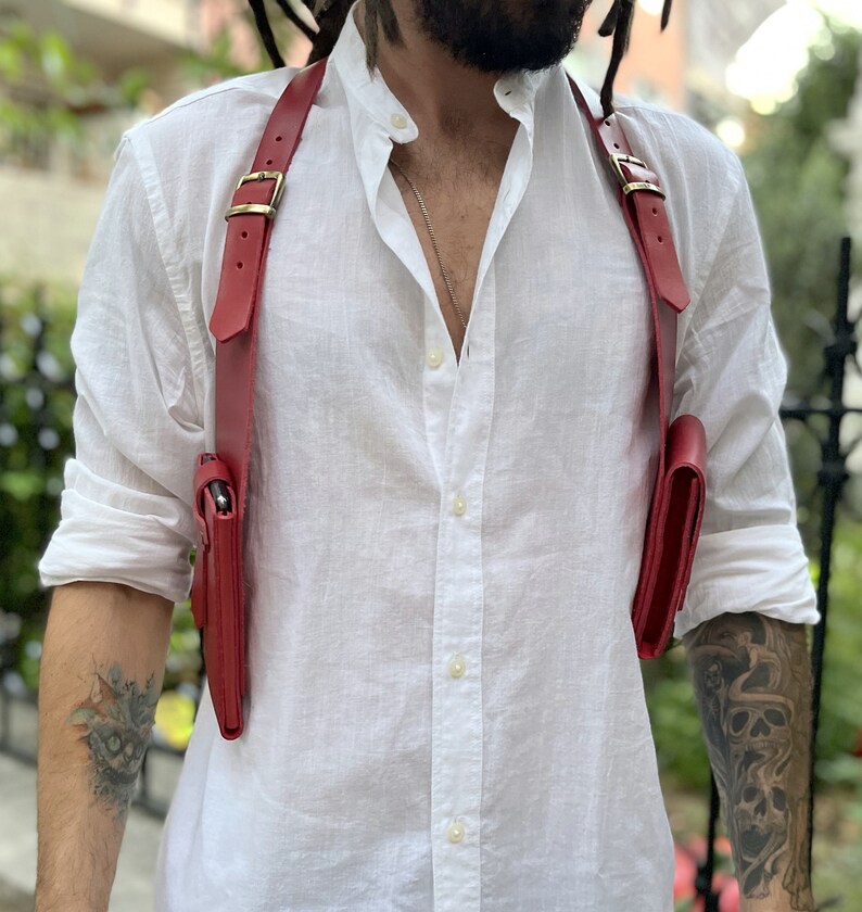 May include: A red leather crossbody bag with two separate compartments. The bag is worn over the shoulder and has a strap that goes across the chest.