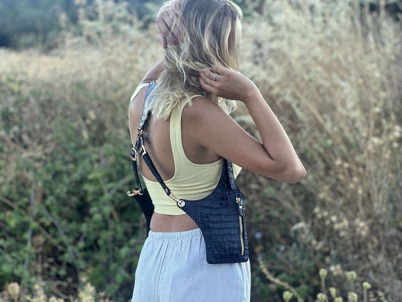 May include: A black leather crossbody bag with a crocodile-embossed design. The bag has a strap that goes over the shoulder and across the chest. The bag has a zipper closure and a small pocket on the front.
