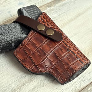 May include: A brown leather holster with a crocodile pattern. The holster has a brown leather strap with two brass snaps.