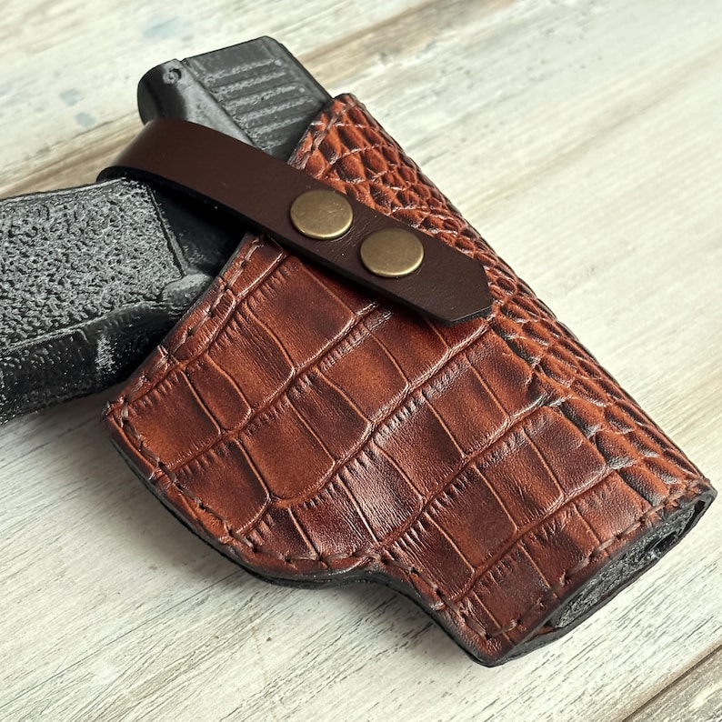 May include: A brown leather holster with a crocodile pattern. The holster has a brown leather strap with two brass snaps.