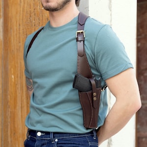 May include: A brown leather shoulder holster with a gun. The holster has adjustable straps and a buckle closure. The gun is black. The person is wearing a teal t-shirt and blue jeans.