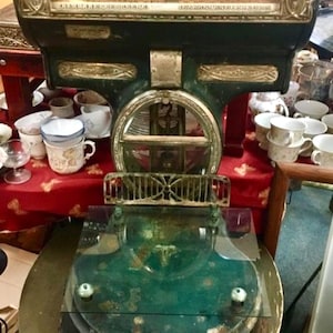 May include: A vintage dark green metal scale featuring a prominent horizontal display window with numerical markings, ornate metallic detailing, and a flat glass weighing platform. The structure includes a circular central frame and a base.