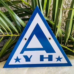 May include: A blue and white triangular sign featuring the letters "A", "Y", and "H" and two stars. The sign has two metal eyelets and is set against a backdrop of green palm leaves.