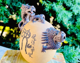 May include: A brown ceramic teapot with two dragon handles. The teapot has Chinese characters on the side. The teapot is sitting on a green and white surface.