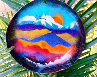 Könnte beinhalten: Ein rundes, handbemaltes Metallkunstwerk, das eine Berglandschaft mit Sonnenuntergang und Vögeln darstellt. Das Kunstwerk weist eine lebendige Farbpalette aus Blau, Orange und Lila mit weißen Berggipfeln auf. Das Stück hat oben ein kleines Loch zum Aufhängen.