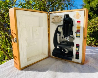 Könnte beinhalten: Ein schwarzes Vintage-Mikroskop mit silberfarbenen Komponenten wird in einem offenen Holzkoffer präsentiert. Der Koffer hat eine hellfarbene Innenausstattung und enthält kleine Fläschchen und Objektträger. Der Koffer ist aus hellfarbenem Holz.