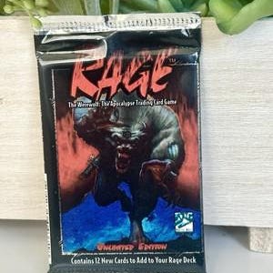 May include: A sealed trading card pack for "Rage: The Werewolf: The Apocalypse Trading Card Game." The pack features a menacing werewolf illustration in red, black, and blue. The text "RAGE" is prominently displayed.