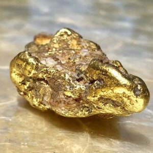 May include: A natural gold nugget with a rough, textured surface. The nugget has a bright, metallic gold color with areas of lighter, possibly quartz, inclusions. The overall shape is irregular and organic, suggesting it was found in its natural state.