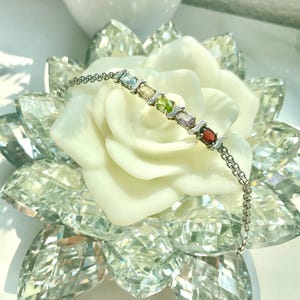 May include: A silver chain bracelet with a row of colorful gemstones. The gemstones are oval-shaped and include blue, yellow, green, purple, and red.