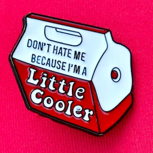 May include: A red, white, and black enamel pin in the shape of a cooler. The pin has the text "DON'T HATE ME BECAUSE I'M A Little Cooler" in white lettering.