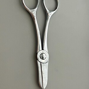 May include: Silver-toned, vintage-style scissors with decorative floral accents on the handles. The scissors have a rounded blade and finger holes. The scissors are on a gray background.