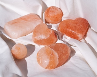Op de afbeelding: Een verzameling roze Himalaya zoutproducten, waaronder hartvormige stenen, bollen en rechthoekige blokken, gerangschikt op een witte stoffen ondergrond. De zoutstukken variëren in grootte en vorm en tonen een natuurlijke, aardse uitstraling.