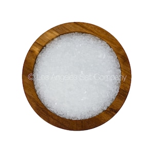 May include: A close-up view of a wooden bowl filled with white sea salt crystals. The bowl is made of light brown wood with a smooth finish. The salt crystals are small and uniform in size, and they are arranged in a dense, even layer.