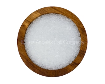 May include: A close-up view of a wooden bowl filled with white sea salt crystals. The bowl is made of light brown wood with a smooth finish. The salt crystals are small and uniform in size, and they are arranged in a dense, even layer.