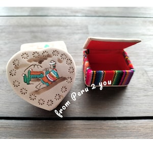 May include: Two small decorative boxes. One box is heart-shaped with a white background and a colourful illustration of a person playing a flute. The other box is square with a red interior and a colourful striped exterior. The text "from Peru 2 you" is written on the image.