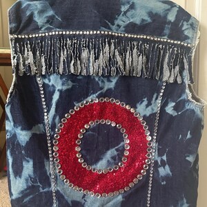 May include: A denim vest with a tie-dye pattern in shades of blue and white. The back features a large red sequined circle, outlined with silver rhinestones. Silver fringe and rhinestones adorn the shoulders and edges.