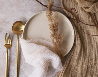 May include: A white ceramic plate with a beige linen runner and gold silverware. The plate is set on a light gray background with a dried branch and dried grass.