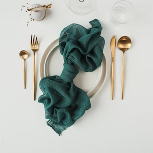 May include: A white plate with a teal green cloth napkin folded in the center. The plate is set with a gold fork, knife, and spoon. There are two clear glass cups and a small vase with dried flowers on a white background.