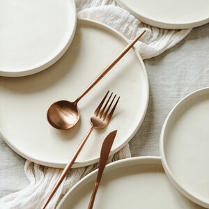 May include: Four white ceramic plates with a rose gold fork, spoon, and knife on a white linen tablecloth.