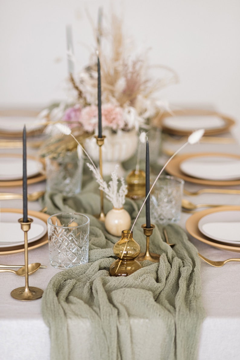 May include: A green table runner with gold candle holders and black candles. There are clear glass vases with dried flowers and a clear glass tumbler on the table.