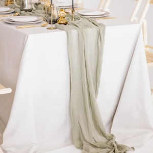 May include: A white tablecloth with a sage green runner draped over it. The runner is gathered at the end of the table and hangs down to the floor. There are gold candlesticks and a vase with dried flowers on the table.