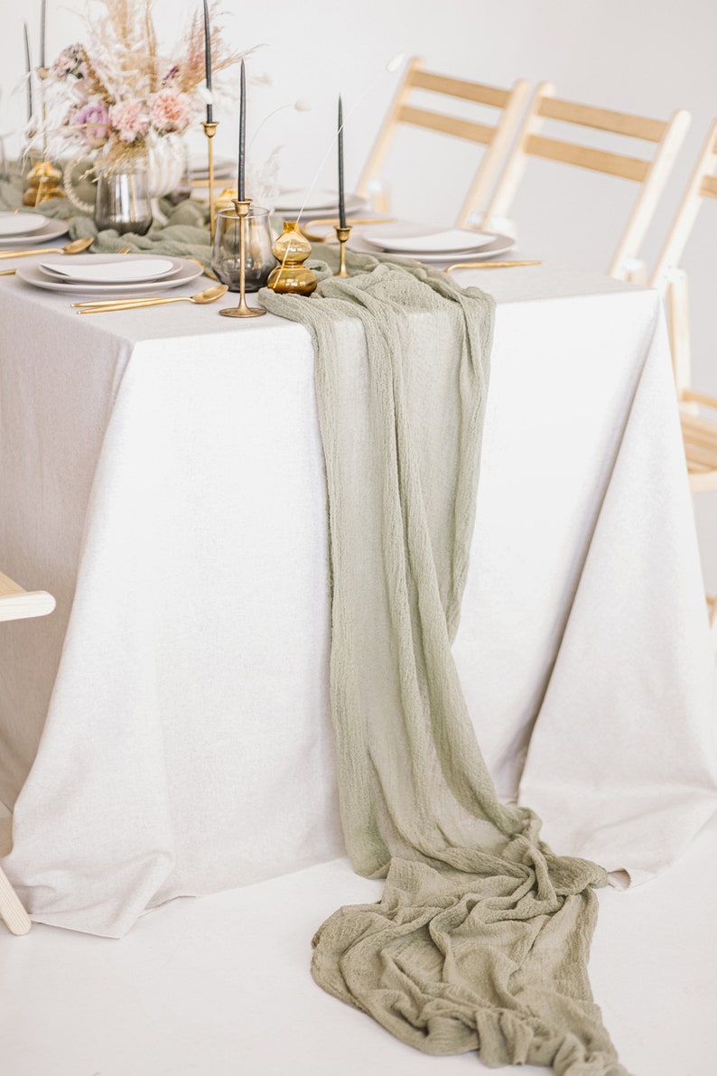 May include: A white tablecloth with a sage green runner draped over it. The runner is gathered at the end of the table and hangs down to the floor. There are gold candlesticks and a vase with dried flowers on the table.
