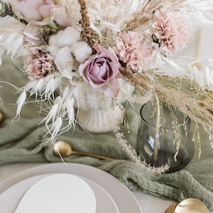 May include: A table setting featuring a white and grey oval plate, gold cutlery, and a green cloth napkin. A vase filled with dried flowers, including pink roses, carnations, and cotton, is positioned in the centre of the table.