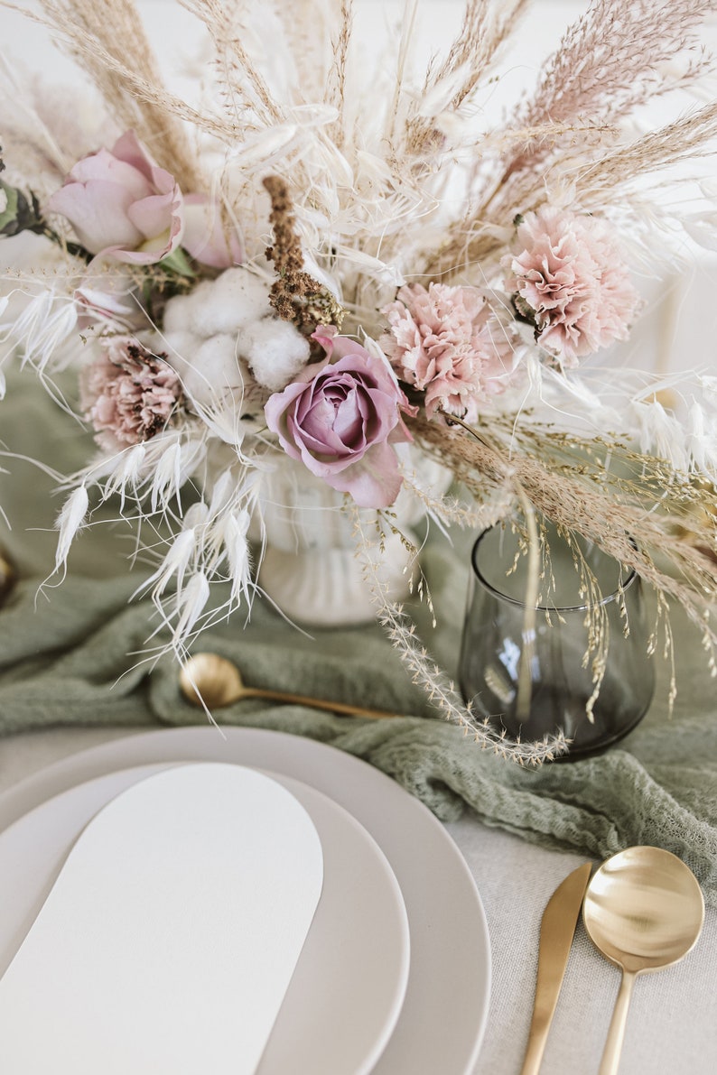 May include: A table setting featuring a white and grey oval plate, gold cutlery, and a green cloth napkin. A vase filled with dried flowers, including pink roses, carnations, and cotton, is positioned in the centre of the table.