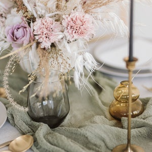 May include: A bouquet of dried flowers in a clear glass vase, with dried pampas grass and other dried foliage. The vase is sitting on a green tablecloth with gold cutlery and a gold candle holder.