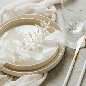 May include: A white ceramic plate with a white ribbon and a dried branch on top. The plate is on a beige linen tablecloth with a glass of water and two silver and gold utensils.