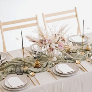 May include: A table setting with a white tablecloth and a green runner. The centrepiece is a bouquet of dried flowers and grasses in a white vase. There are gold candlesticks, plates, and cutlery on the table.