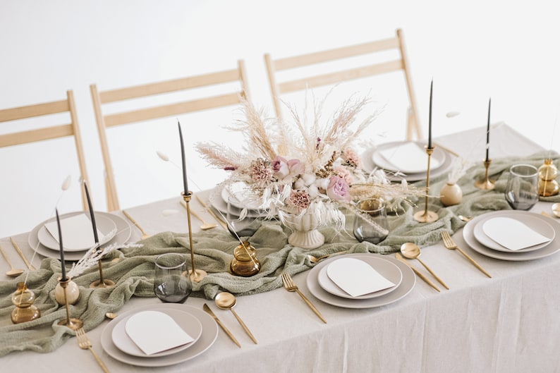 May include: A table setting with a white tablecloth and a green runner. The centrepiece is a bouquet of dried flowers and grasses in a white vase. There are gold candlesticks, plates, and cutlery on the table.