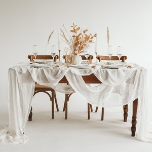 May include: A white tablecloth with a delicate, gauzy texture drapes over a wooden table set for a dinner party.  The table is decorated with a vase of dried flowers and greenery, and four place settings with clear glass goblets and white plates.