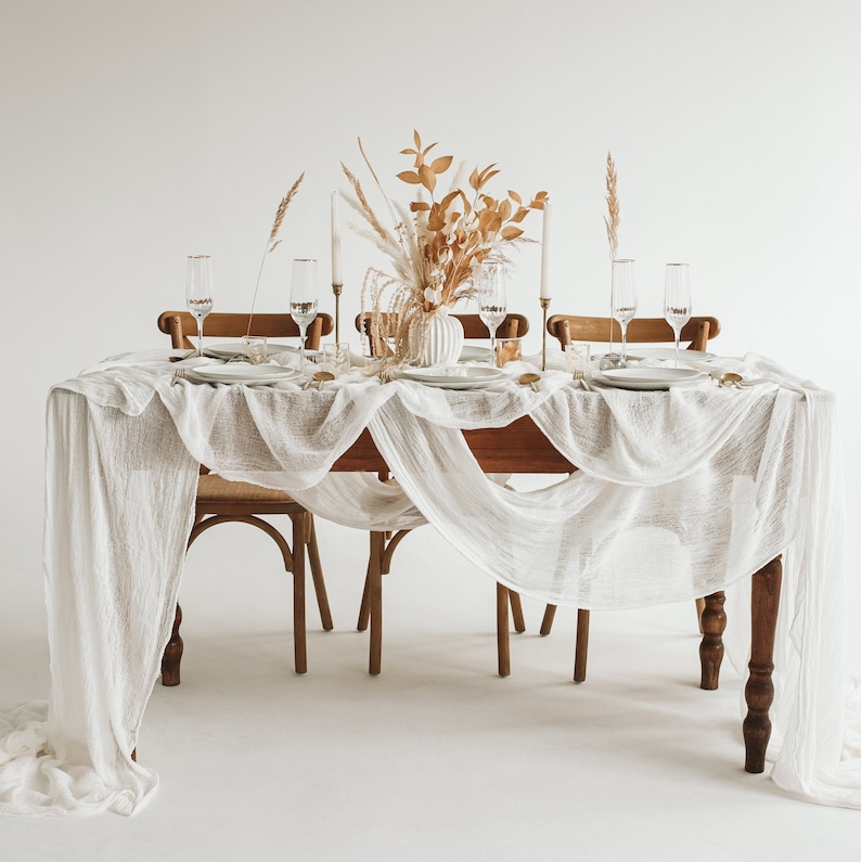May include: A white tablecloth with a delicate, gauzy texture drapes over a wooden table set for a dinner party.  The table is decorated with a vase of dried flowers and greenery, and four place settings with clear glass goblets and white plates.