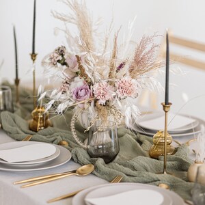 May include: A table setting with a green tablecloth and white plates. A bouquet of dried flowers in a clear glass vase is the centrepiece. The flowers are pink, purple, and white. There are gold candlesticks and gold vases on the table.