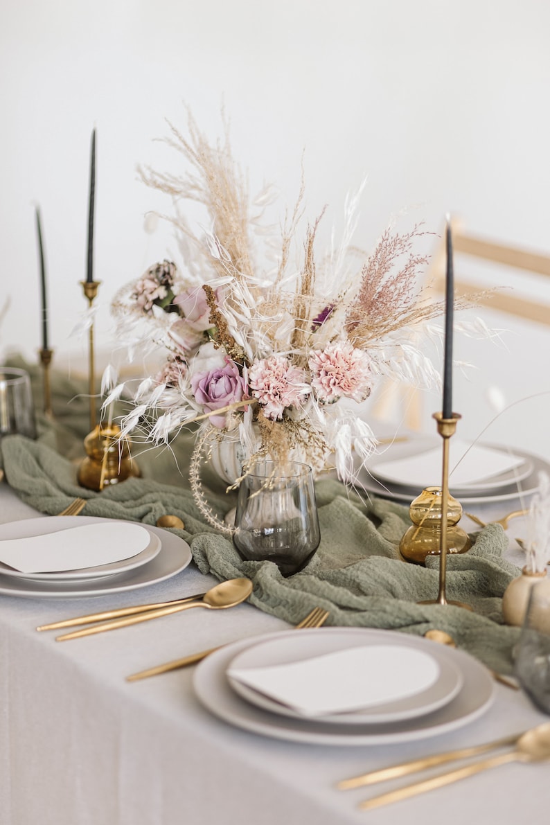 May include: A table setting with a green tablecloth and white plates. A bouquet of dried flowers in a clear glass vase is the centrepiece. The flowers are pink, purple, and white. There are gold candlesticks and gold vases on the table.