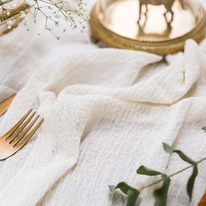 Puede incluir: Un mantel de lino blanco con un tenedor y un cuchillo dorados, una rama de vegetación y una pequeña cesta tejida en el fondo.