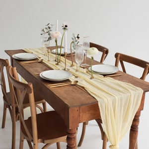 May include: A rustic wooden table set with a yellow linen table runner, white plates, gold cutlery, and white flowers in gold vases. Two wooden chairs are placed on either side of the table.