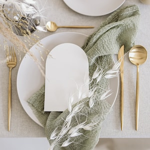May include: A white card with an arched top sits on a light grey plate. A green cloth napkin with white dried flowers is placed on the plate. Gold silverware and a gold spoon are on the table.