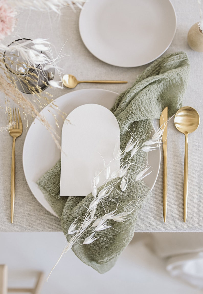 May include: A white card with an arched top sits on a light grey plate. A green cloth napkin with white dried flowers is placed on the plate. Gold silverware and a gold spoon are on the table.