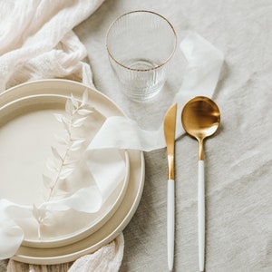 May include: A white linen tablecloth with a white ceramic plate, a glass, a gold spoon, and a gold knife. The plate has a white ribbon and a sprig of white leaves on top.