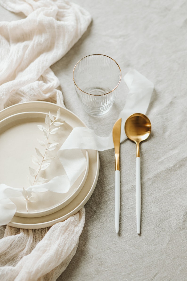 May include: A white linen tablecloth with a white ceramic plate, a glass, a gold spoon, and a gold knife. The plate has a white ribbon and a sprig of white leaves on top.