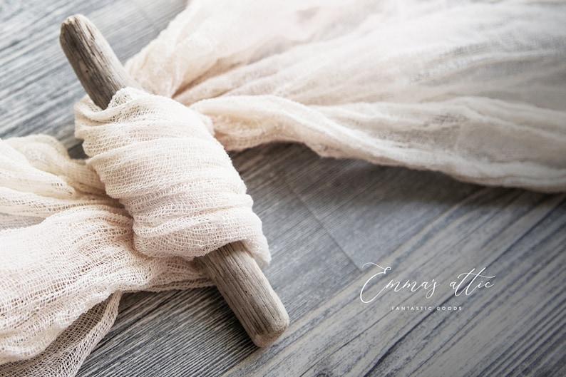 May include: A piece of white gauze fabric draped over a piece of driftwood. The fabric is textured and has a natural, rustic look. The driftwood is light brown and has a rough surface. The fabric and driftwood are on a gray wooden surface. The text "Emma's Attic Fantastic Goods" is visible in the lower right corner of the image.