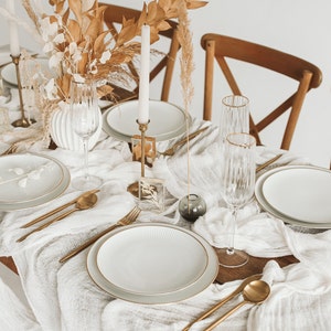 May include: A white tablecloth with a gold rimmed plate, gold silverware, and two champagne flutes. Dried flowers and a candle are in the background.