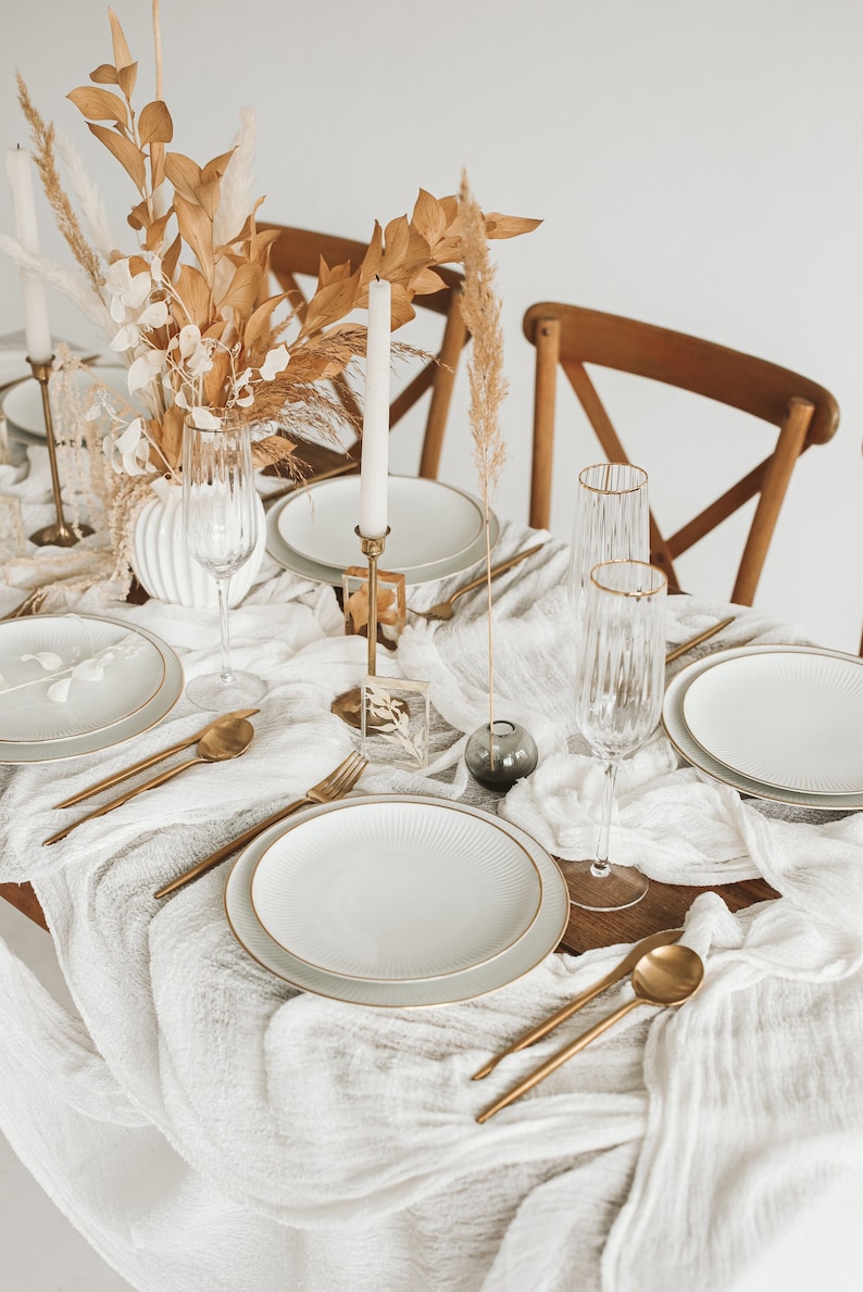 May include: A white tablecloth with a gold rimmed plate, gold silverware, and two champagne flutes. Dried flowers and a candle are in the background.