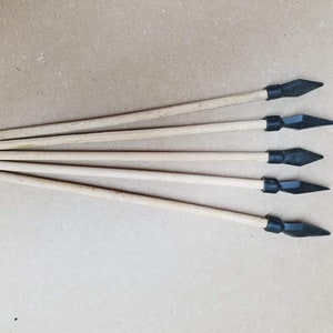Könnte beinhalten: Fünf Holzpfeile mit schwarzen Pfeilspitzen. Die Pfeile haben hellbraune Schäfte und schwarze Spitzen. Die Pfeilspitzen sind mit einem schwarzen Band befestigt. Die Pfeile sind diagonal auf einer hellbraunen Oberfläche angeordnet.