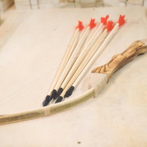 May include: A wooden bow and six arrows with red fletching and black tips. The bow is made of wood and has a brown leather grip.
