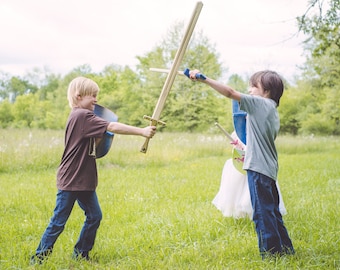 May include: Two young boys wearing casual clothing are playing with toy swords and shields in a grassy field. One boy is wearing a brown shirt and blue jeans, while the other is wearing a gray shirt and blue jeans. They are both holding wooden swords and shields, and they are pretending to fight each other.